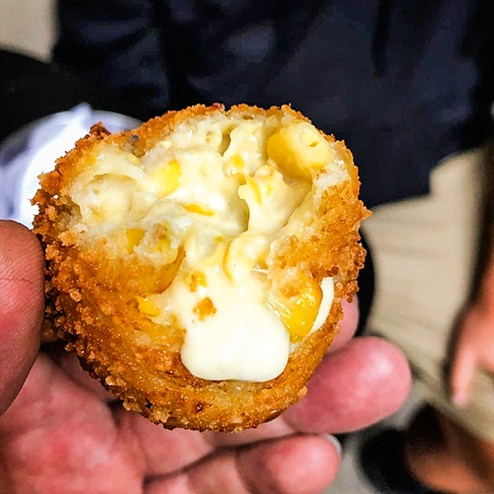 Bolinho – Moela Bolinho de milho, com queijo gorgonzola derretido. Na imagem aparece os dedos da pessoa segurando o bolinho.