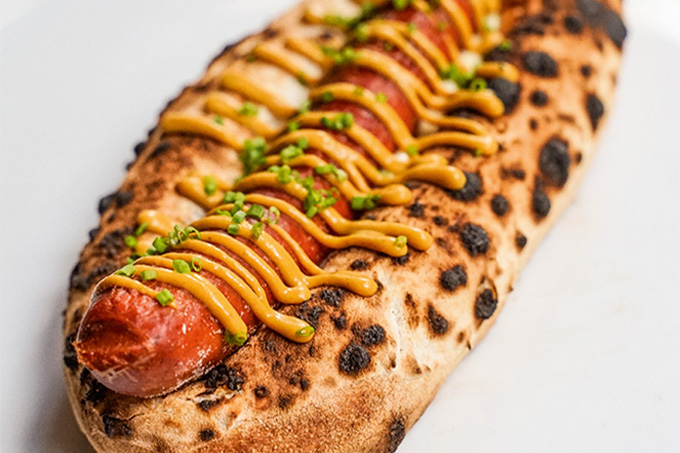 Foto da pizza de cachorro-quente da Deveras Pizza, com salsicha coberta de mostarda sobre massa de pizza esticada, em fundo branco.