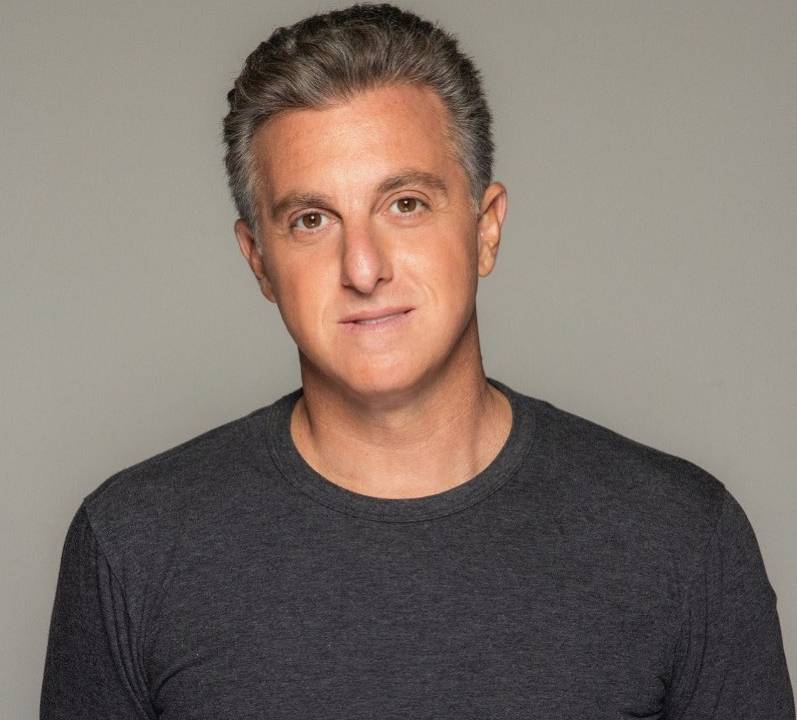 Imagem mostra foto do apresentador Luciano Huck. Ele usa uma camiseta e olha com um leve sorriso para a câmera