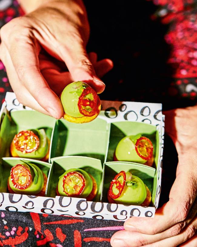 Foto de seis bombons de guacamole em caixa para delivery com divisão para cada um e uma mão oferecendo um deles.