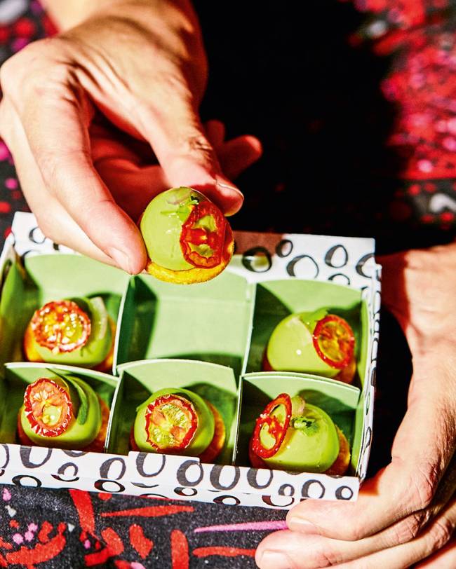 iFood Gourmet – Maní Foto de seis bombons de guacamole em caixa para delivery com divisão para cada um e uma mão oferecendo um deles.