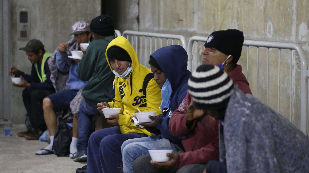 Imagem mostra pessoas com gorros e agasalhos, sentadas uma ao lado da outra, com embalagens de isopor, tomando sopas; eles estão no metrô