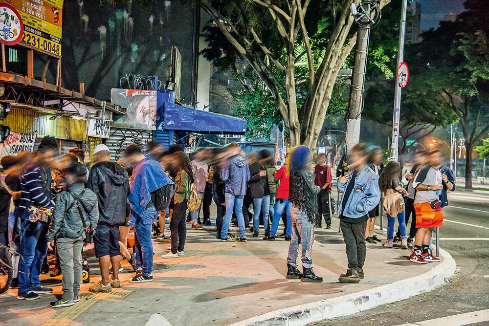 A imagem mostra uma esquina repleta de jovens, sem máscaras, próximo a um bar