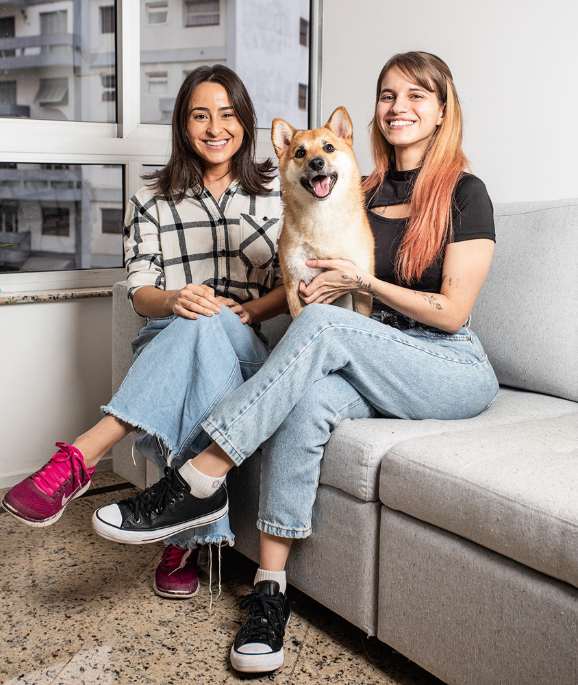 Moradia compartilhada A imagem mostra as duas amigas sentadas em um sofá, com o cachorro no colo, sorrindo para a câmera.