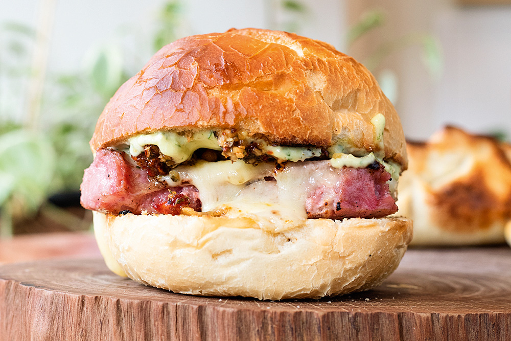 Sanduíche choripán sobre tampo de madeira, em pão redondo similar ao francês recheado de linguiça, queijo e maionese verde.