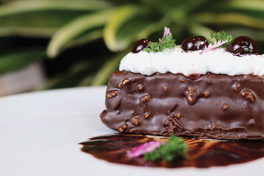 Dalva e Dito – Bolo floresta negra Bolo floresta negra, feito à base de pão de ló, com recheio de brigadeiro com chantili, cereja e puxuri, servido em uma prato raso de cor branca