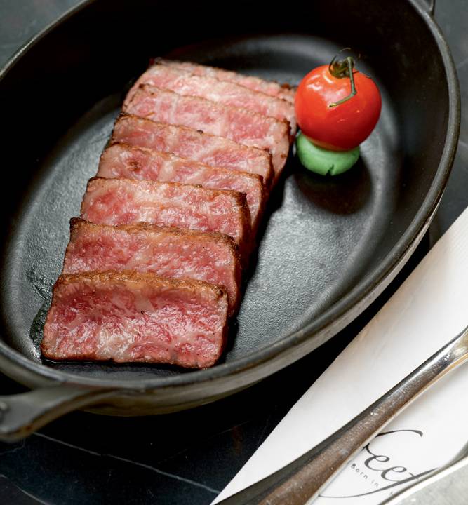 Panelinha oval de metal preta com oito fatias de carne colocadas parcialmente uma sobre a outra ao lado de tomatinho cereja sobre disco de wasabi.