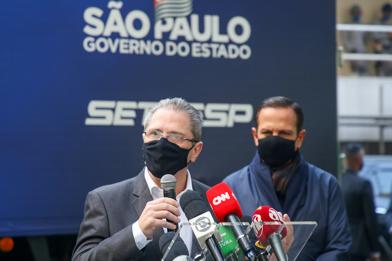 A imagem mostra Jean com um microfone falando em frente a microfones de vários veículos de comunicação. Atrás, ao fundo e desfocado, está João Doria, o observando.