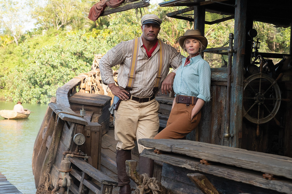 A imagem mostra Dwayne Johnson e Emily Blunt apoiados em um barco, em um rio cercado por mata. Eles vestem roupas de época, de dois expedidores, usando chapéus.
