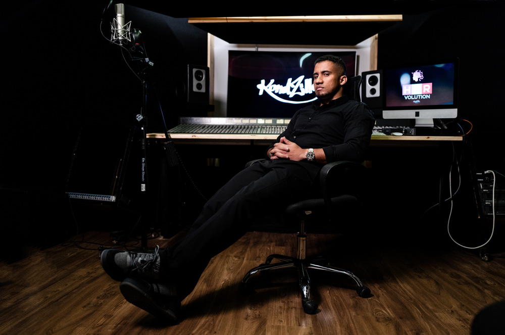 A imagem mostra Konrad, sentado em uma cadeira dentro de um estúdio. Há, ao fundo, um enorme mesa de mixagem de som e um telão ao fundo escrito KondZilla.