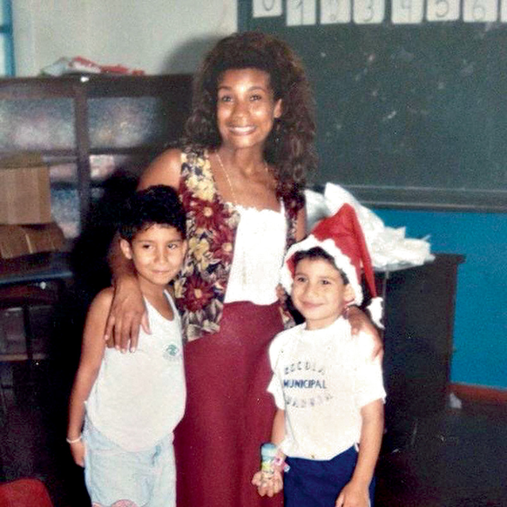 Konrad-Criança-com-a-Mãe A imagem mostra a mãe de Konrad, Áurea Cunha, com mão nos ombros de duas crianças, com os três sorrindo para a câmera. Ao fundo, há uma lousa verde mostrando que eles estão em uma sala de aula.