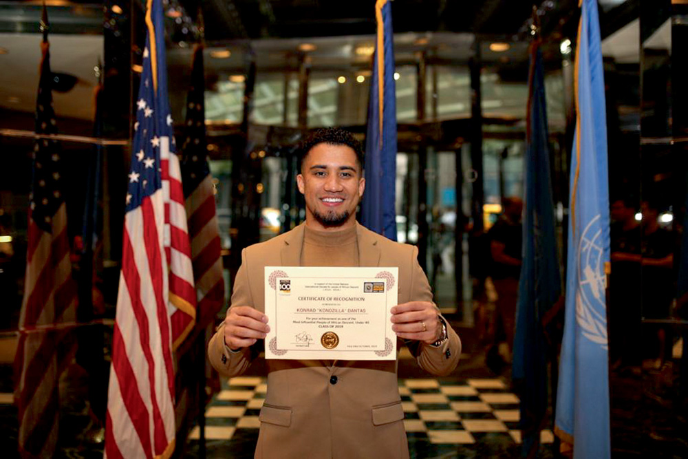 Konrad-Honra-Internacional A imagem mostra Konrad segurando o certificado e ao fundo há as bandeiras dos Estados Unidos e ONU.