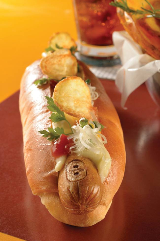 Lanchonete da Cidade Cachorro-quente coberto por mostarda, salsinha e batata chips sobre bancada avermelhada.