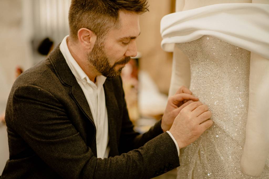 Lucas Anderi – Disney Princess Wedding Collection (Jeff Porto) (3) Lucas Anderi costura trecho de vestido de noiva na imagem. Aparece de paletó preto e camisa branca.