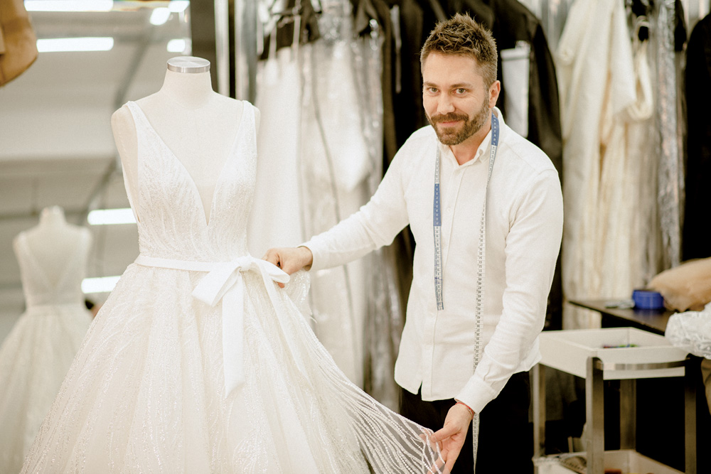 A imagem mostra Lucas puxando levemente um vestido segurado por um manequim. Ele está com uma fita métrica sobre os ombros e sorrindo para a câmera.