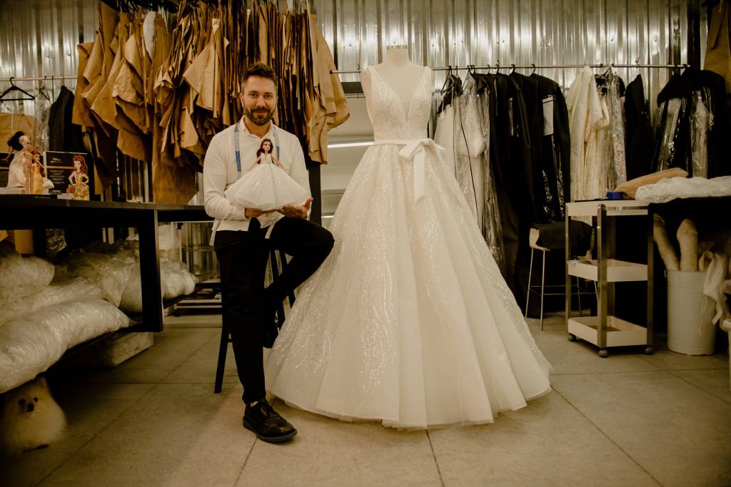 Lucas Anderi aparece sentado ao lado de vestido de noiva. Veste calça preta e camisa branca, com fita métrica no pescoço e segurando boneca.