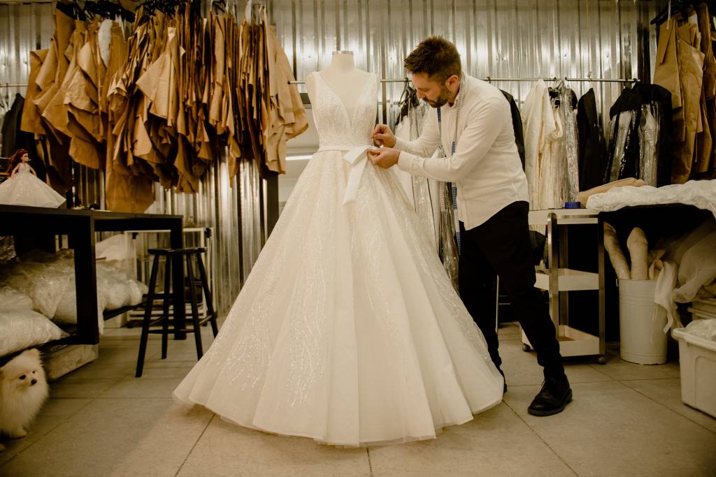 Lucas Anderi – Disney Princess Wedding Collection (Jeff Porto) (6) Lucas Anderi costura trecho de vestido de noiva na imagem. Aparece de paletó preto e camisa branca ao lado do vestido em seu ateliê.