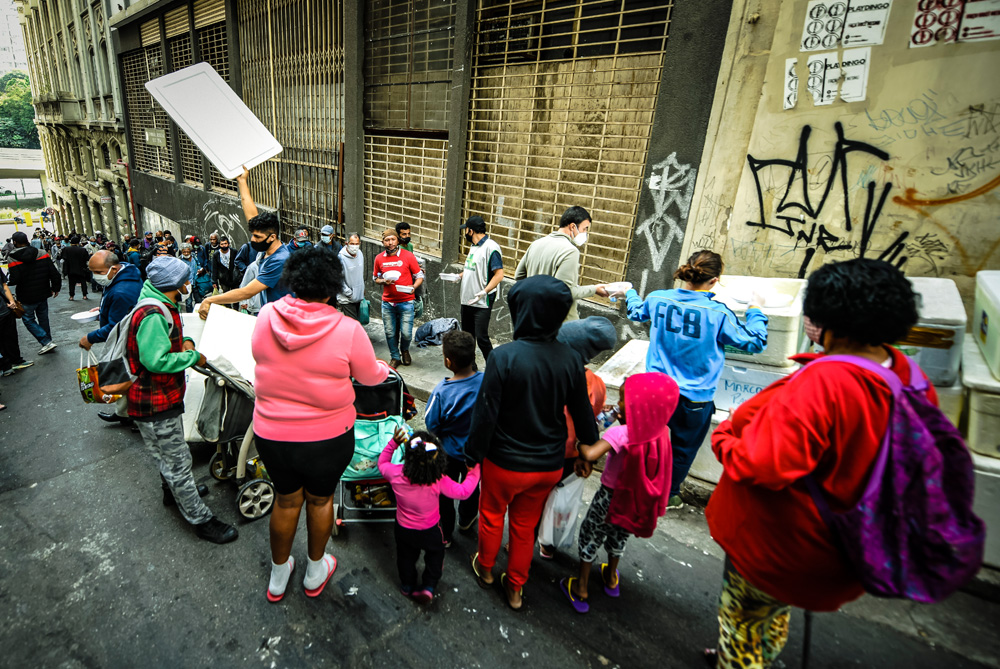 A imagem mostra uma fila de pessoas na rua em frente de grandes isopores onde há pessoas retirando marmitas de dentro.