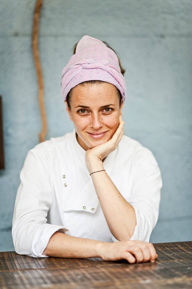 Chef Helena Rizzo sentada atrás de mesa vestida com dólmã e lenço lilás no cabelo, com o cotovelo apoiado sobre a mesa e a mão segurando a bochecha.