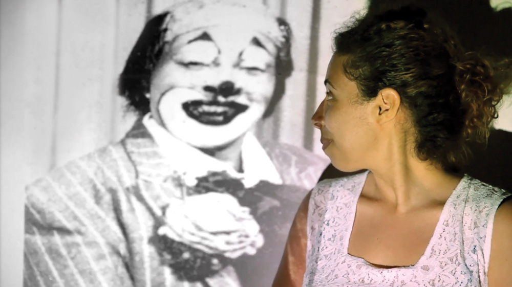 Mariana-avó-palhaça-história-dia-dos-avós.jpg mariana de perfil olhando para tras, onde está sendo projetado o documentário de sua avó, com uma cena em que sua avó aparece vestida de palhaço