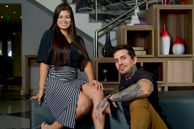 arthur e luana na sala de casa posando para a foto. eles estão na sala, luana está sentada no sofá com um braço no sofá e outro no joelho. arthur está a seu lado sentado no tapete. tem uma escada ao fundo