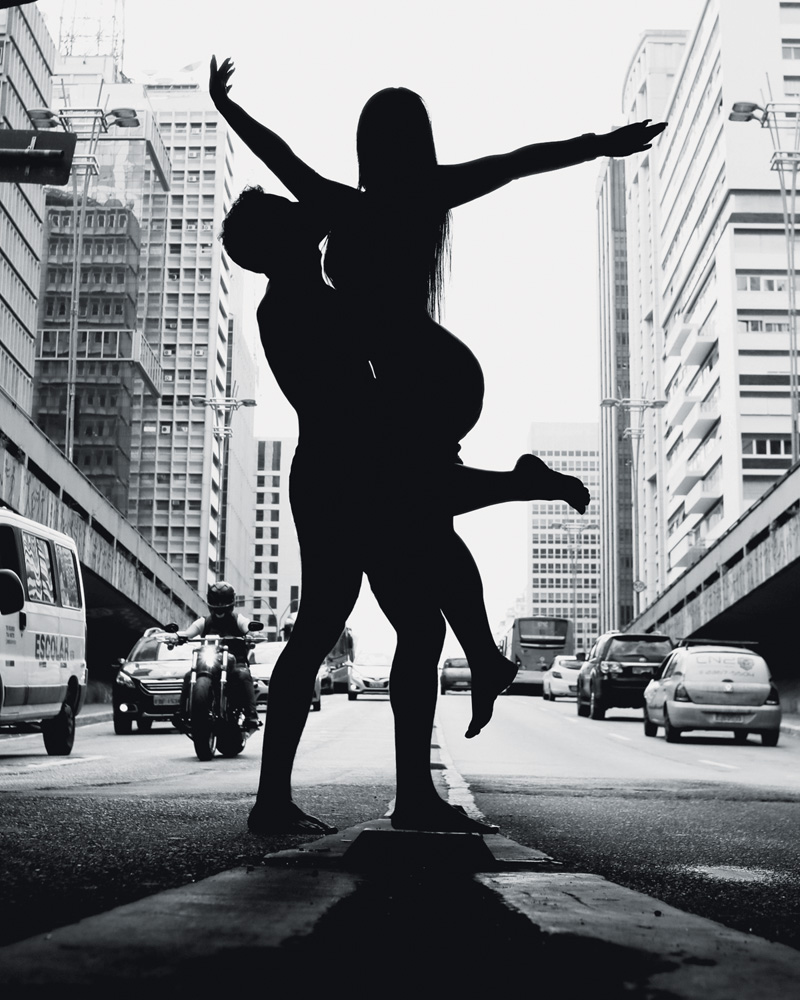 Nosso-Louco-Amor-Arthur-Luana-amor-livre-swing-ensaio-paulista.jpg.jpg foto em preto e branco da silhueta de arthur e luana posando nus para a foto na paulista. eles estão no meio da rua, com a avenida ao fundo, fazendo uma pose em que ele a segura, enquanto ela abre os braços