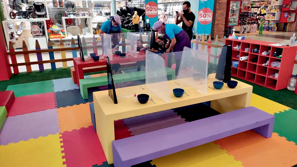 Dentro de um shopping, há um cercadinho, com tapetes de tatame e mesas para crianças com materiais de desenho