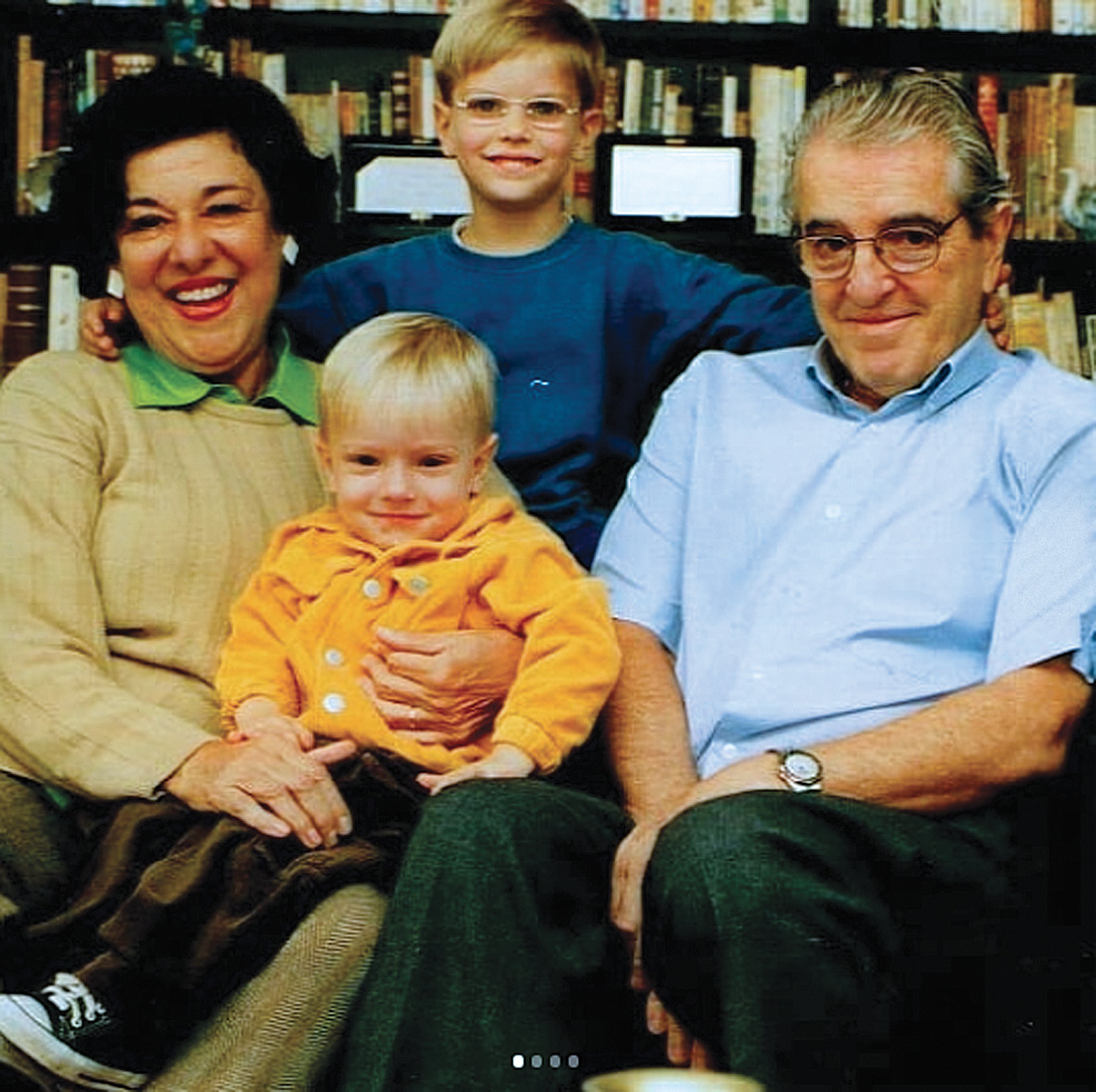 Ruth-Rocha-história-avós-dia-dos-avós.jpg ruth rocha com seu marido, eduardo rocha, e seus netos pedro e miguel, sorrindo posando para a foto. pedro está sentado no colo de ruth, que está sentada ao lado de eduardo. miguel está de pé atrás deles com um braço nas costas de cada avô