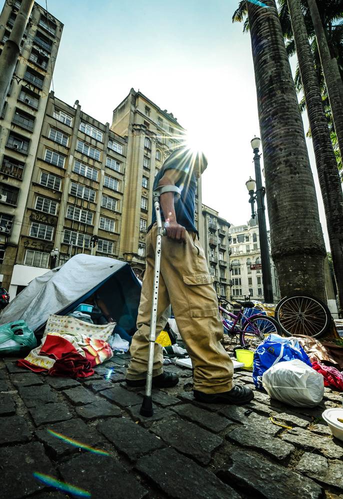 A imagem mostra uma foto, vinda do chão, observando Paulo, muleta, andando. No fundo, há uma barraca na calçada e itens espalhados pelo chão.