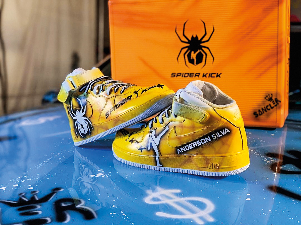 Sancler-artista-tênis-japão-anderson-silva.jpg par de tênis do anderson silva com estampa de aranha e o nome do lutador e 'spider', seu apelido. atrás, um pano amarelo com a estampa de aranha