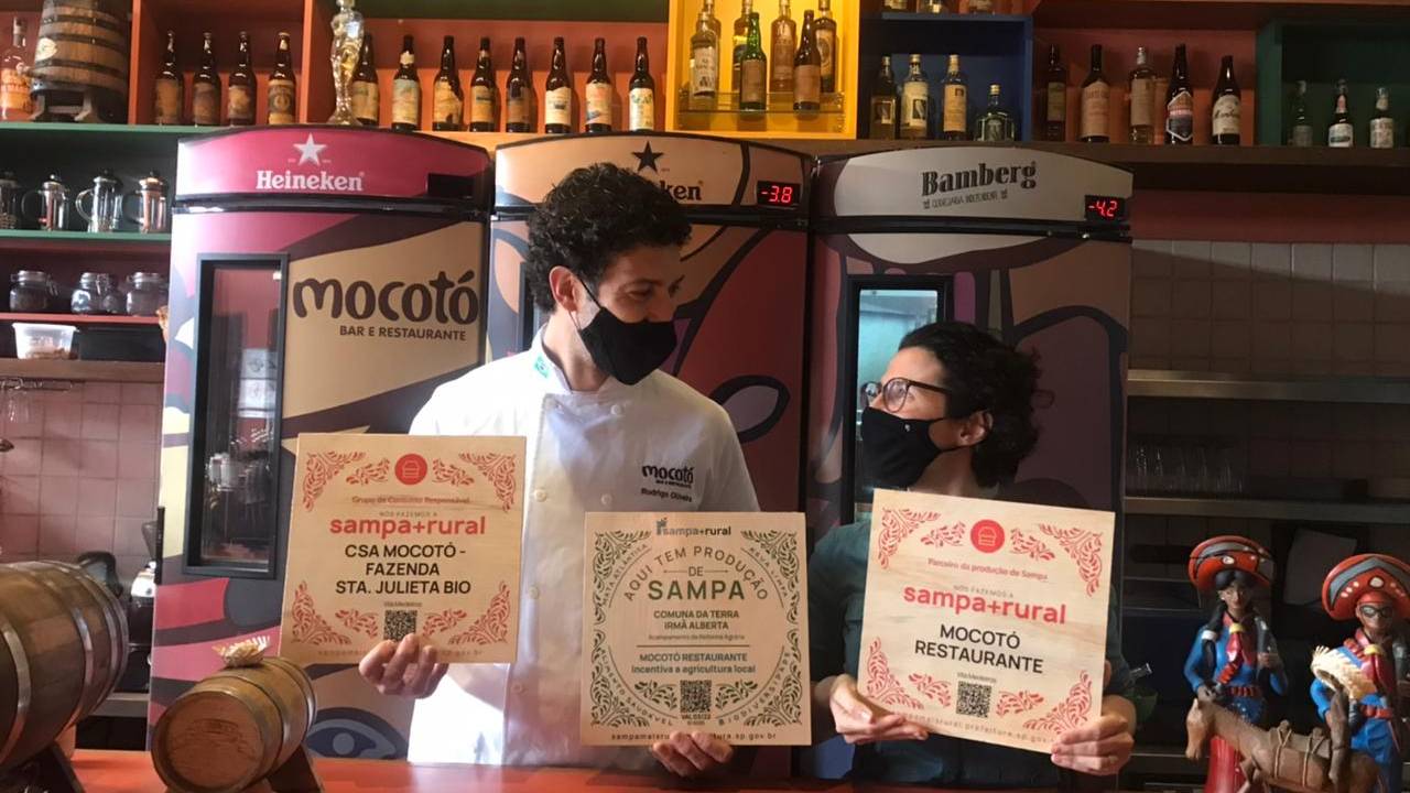 Rodrigo Oliveira e Adriana Salay de máscara segurando placa do Sampa+Rural no balcão de bar do Mocotó.