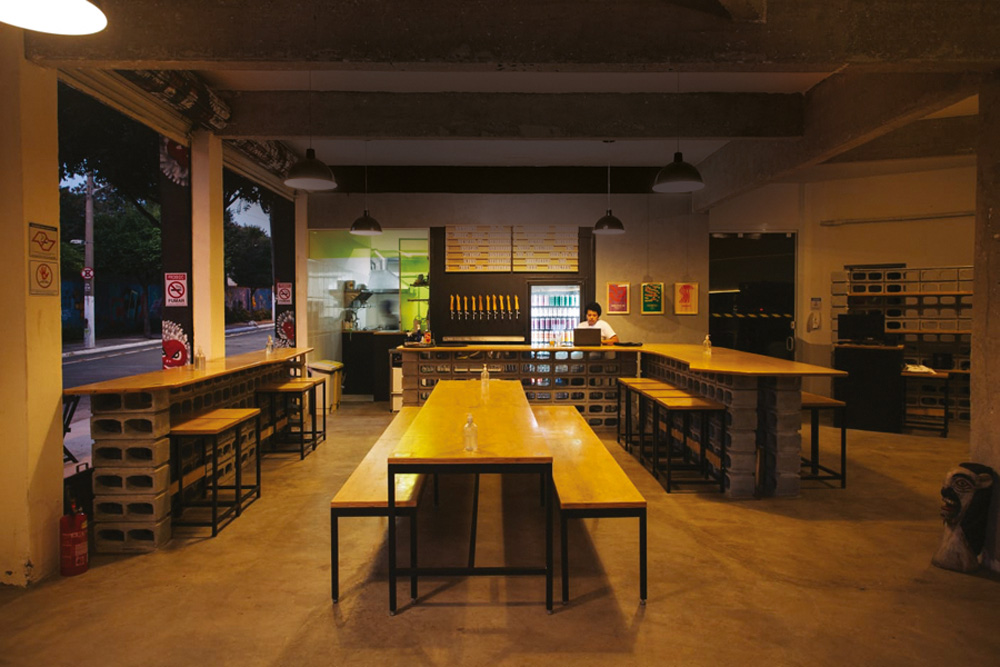 Abertura Gastronomia – Cervejaria Central Salão da Cervejaria Central na Barra Funda, em meia luz com mesas e bancos longos de madeira ao centro, direita e esquerda.