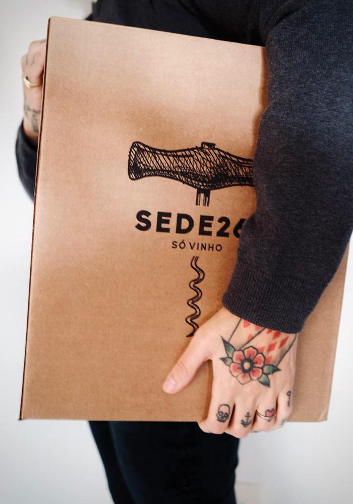 Sede261 Caixa de papelão com o logo do Sede261 é segurada por pessoa de lado com tatuagens na mão.