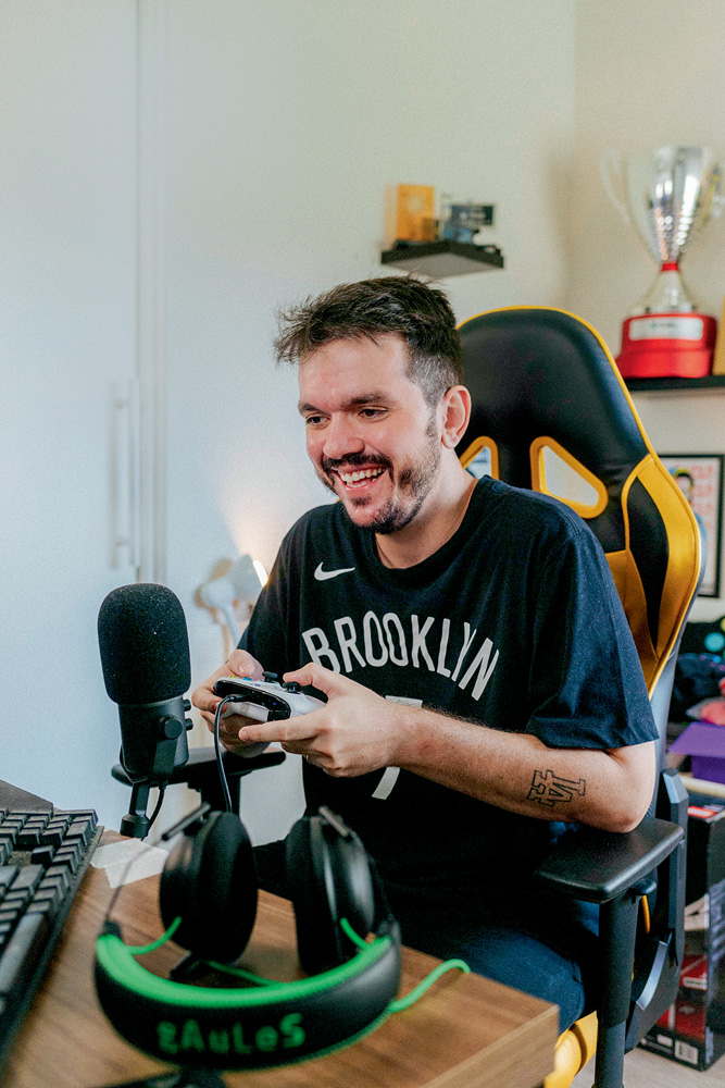 O ex-jogador profissional Gaules: segundo streamer mais visto do mundo A imagem mostra Gaules, sentado em uma cadeira gamer, rindo para seu monitor e segurando um controle de videogame. Há um microfone profissional na sua frente também.