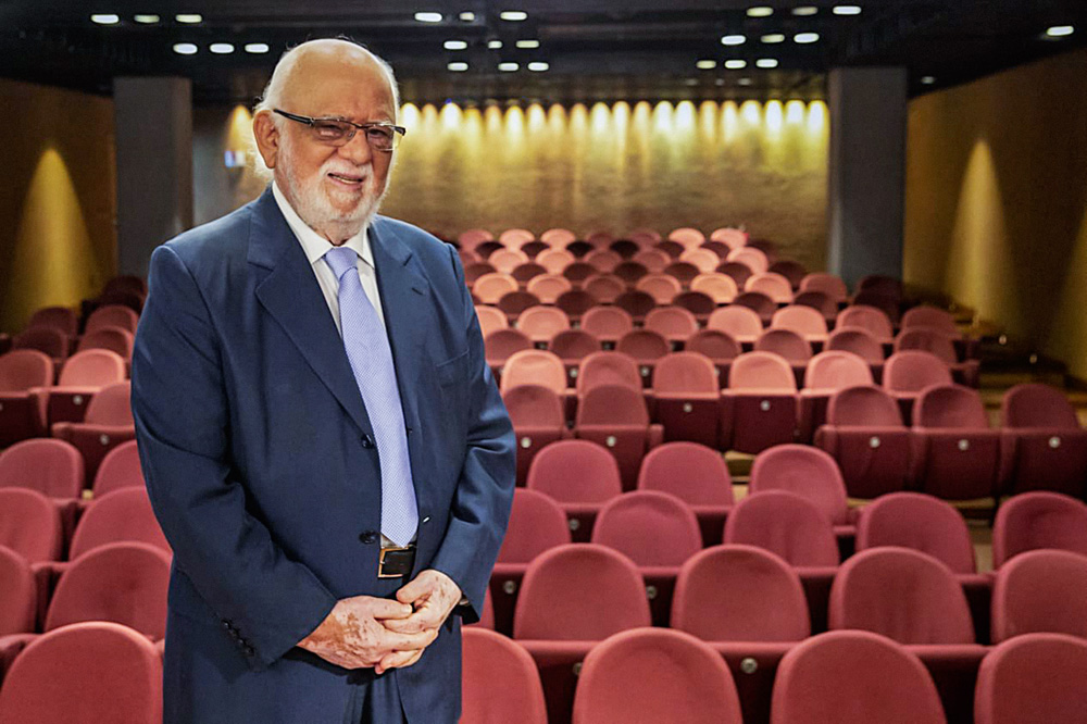 A imagem mostra Danilo em cima de um palco. No fundo, é possível ver cadeiras vazias de um teatro.