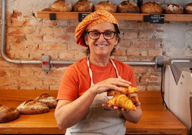 claudia rezende – zestzing Mulher com avental e pães na mão possa em frente a prateleiras de pães