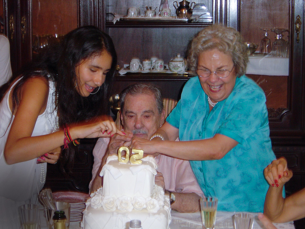 gabriela-del-moro-bolo-isopor-história-dia-dos-avós.jpg gabriela junto de seus avós, lúcio e elvira, com o bolo dos avós na frente. eles estão cortando o bolo, com as mãos juntas na faca. gabriela e elvira estão de pé e sorriem, enquanto lúcio está com expressão fechada