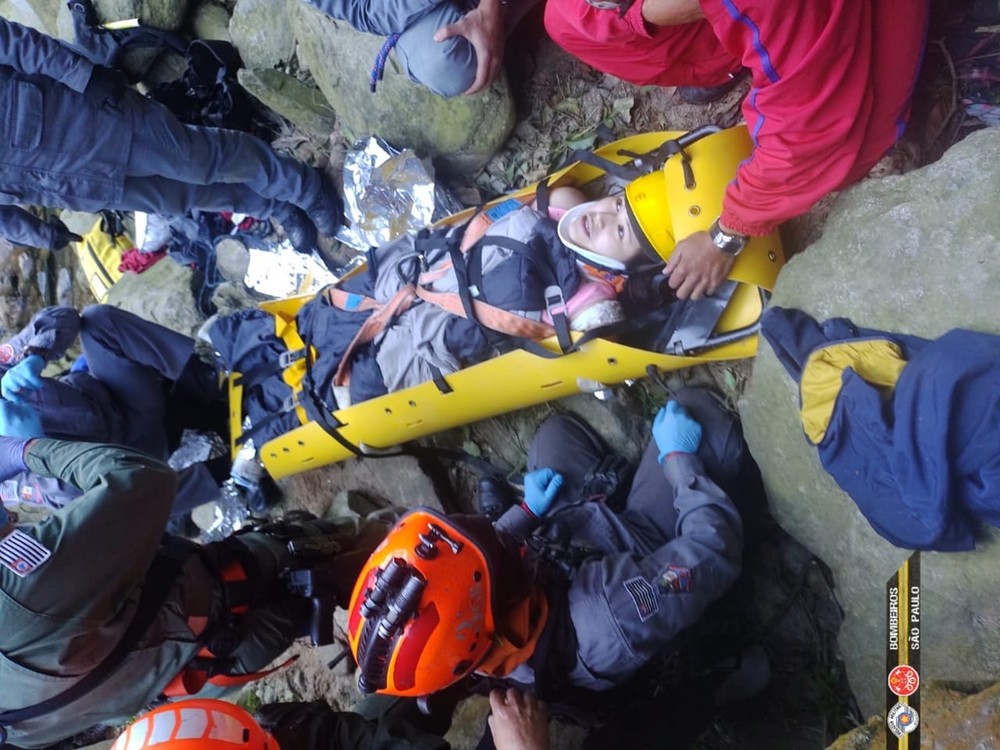 foto tirada de cima para baixo de mulher que sofreu a queda. ela está em uma maca amarela dos bombeiros e está em volta de diversos agentes