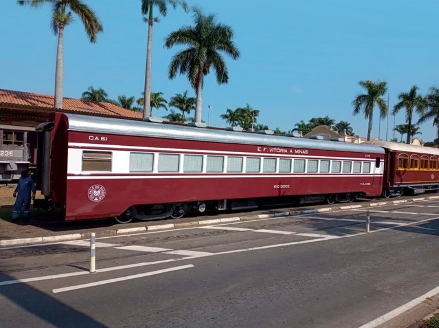 Imagem mostra vagão antigo reformado e estacionado em estação de trem. A pintura tem cor vinho e detalhes em alumínio. Ao fundo, aparecem palmeiras e um céu bem azulado.