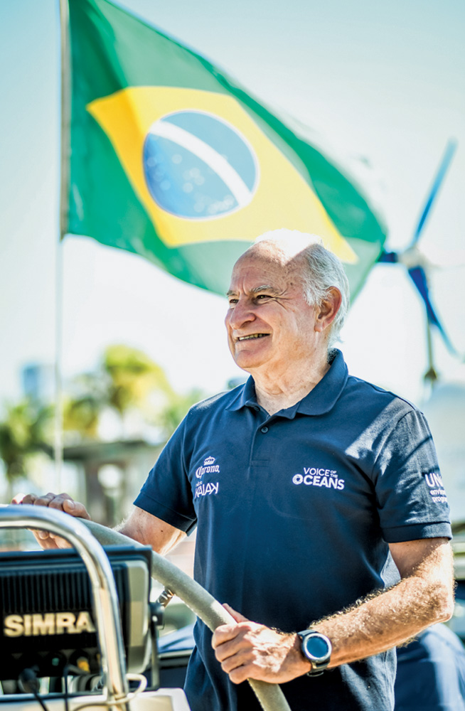 vilfredo-familia-schurmann-nova-aventura-poluicao-oceanos-historias-navegacao.JPG.jpg vilfredo sorrindo olhando para o horizonte segurando o voltante do barco Kat. ao fundo, tem uma bandeira do brasil balançando ao vento