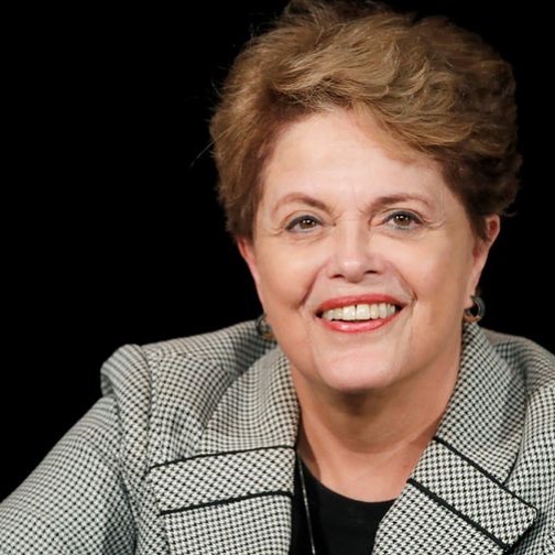 Imagem de Dilma sorrindo. O fundo da foto é preto. Ela veste uma casaco cinza.