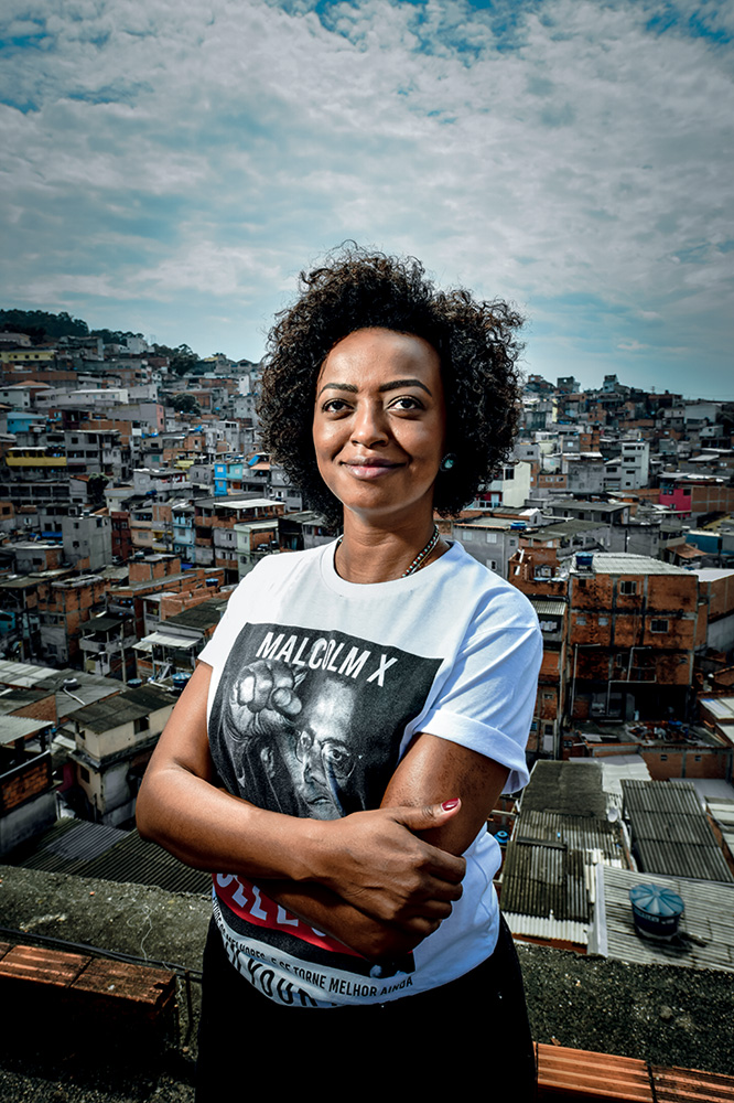 Imagem mostra Aline Torres de braços cruzados. A fundo, comunidade da capital paulista