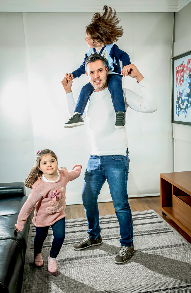 EDUARDOVERISSIMO_6.JPG A imagem mostra Eduardo, sorrindo, com Vitor sobre seus ombros, de cavalinho, e Julia correndo em volta dos dois em uma sala.