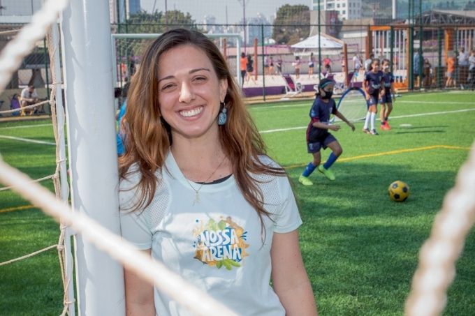 foto de julia vergueiro de trás de barbante do gol. ela está encostada em trave de gol e com camisa do nossa arena. ao fundo, mulheres estão jogando futebol utilizando máscaras