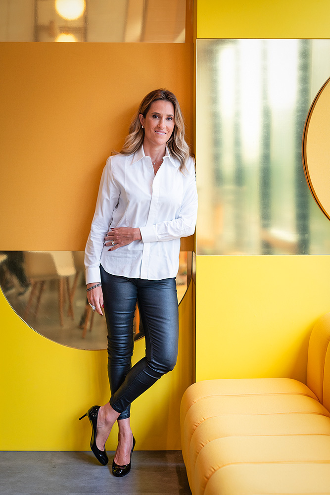 Karla, Cimed (Naira Mattia) Karla Marques Felmanas posa de pé e encostada em parede. Todo o cenário é amarelo-canário e inclui um sofá à direita. Ela veste calça preta e camisa branca. Está com uma das pernas dobradas e um dos braços encostados na barriga.