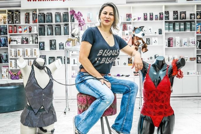 maísa pacheco sentada em banqueta estofada posando para a foto em sua sex shop e ao lado de dois manequins vestidos de lingeries. seu corpo está um pouco de lado e ela está apoiada em um dos manequins