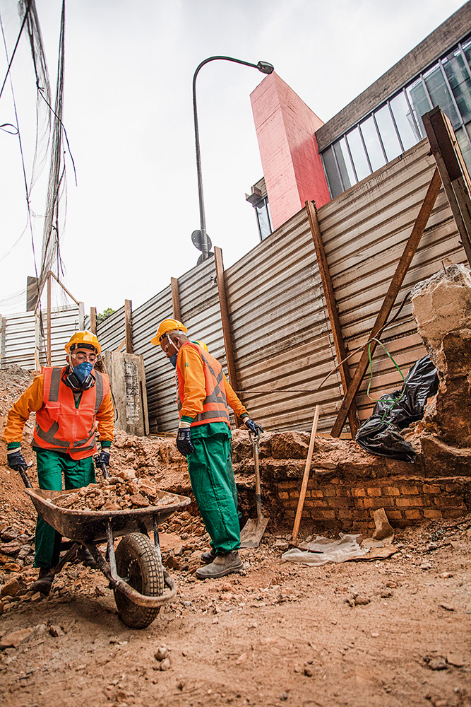 MASP A imagem mostra um canteiro de obras, com dois funcionários trabalhando em meio a terra. Ao fundo, é possível ver uma parte do Masp acima de um tapume do canteiro.