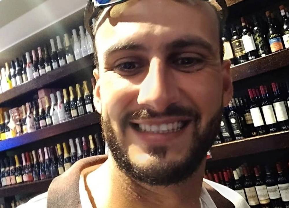 A imagem mostra uma selfie de Matheus. Ele está com um óculos na testa, sorrindo para a câmera, em uma setor de vinho de um supermercado.