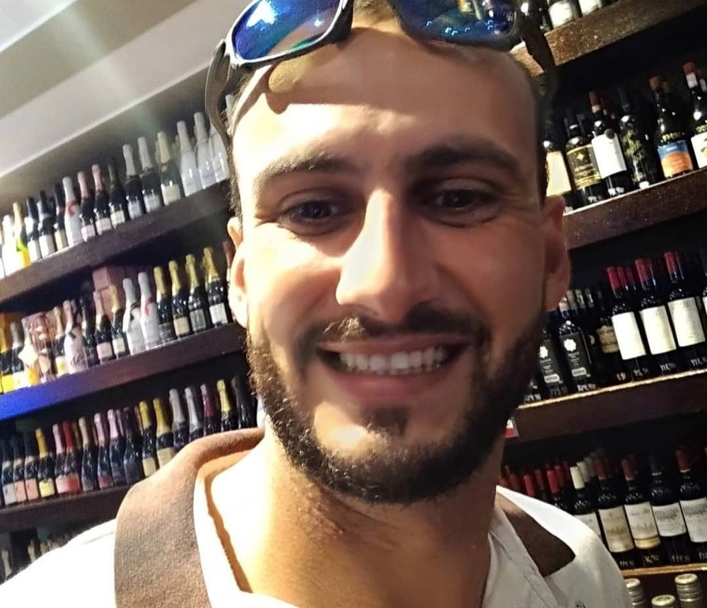 A imagem mostra uma selfie de Matheus. Ele está com um óculos na testa, sorrindo para a câmera, em uma setor de vinho de um supermercado.