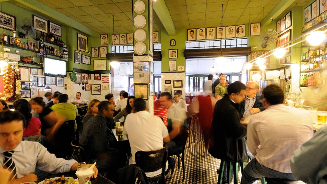 Bar com pessoas ocupando mesas e banquetas altas do balcão